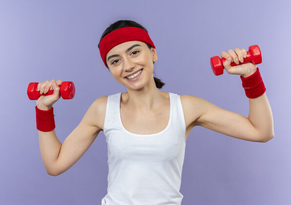 人身穿运动服 头箍 双手举着两个哑铃的年轻健身女士站在紫色的墙壁上愉快地微笑着做运动姿势健身女人