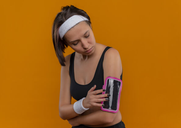 人身穿运动服的年轻健身女士 头戴着智能手机袖标 站在橙色的墙上移动健身头带