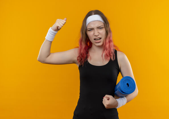 健身身穿运动服的年轻健身女士手持瑜伽垫举起拳头兴奋而快乐地站在橙色的墙上提高女人瑜伽