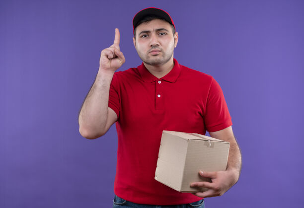 人年轻的送货员穿着红色制服 拿着帽子的盒子 用食指指着紫色的墙上 脸上带着严肃的表情递送目录严肃