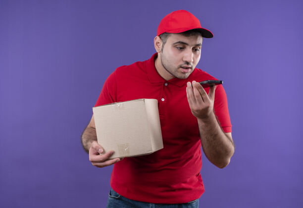 手持年轻的送货员穿着红色制服 戴着帽子拿着一个盒子 站在紫色的墙上用他的智能手机发送语音信息年轻人制服帽子