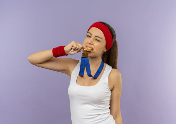 运动员一位身穿运动服 头上戴着金牌的头带 咬着奖牌的年轻健身女士站在灰色的墙上 看上去很自信站人运动
