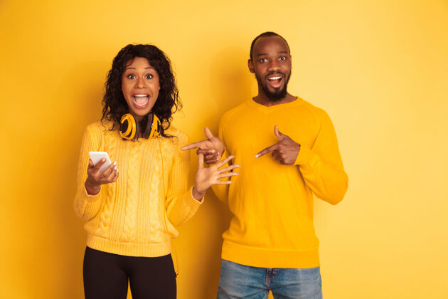 夫妇年轻感性的非裔美国人 穿着黄色背景的明亮休闲服美丽的情侣人类情感的概念 面部表情 关系在智能手机上震惊地指着面部年轻时尚