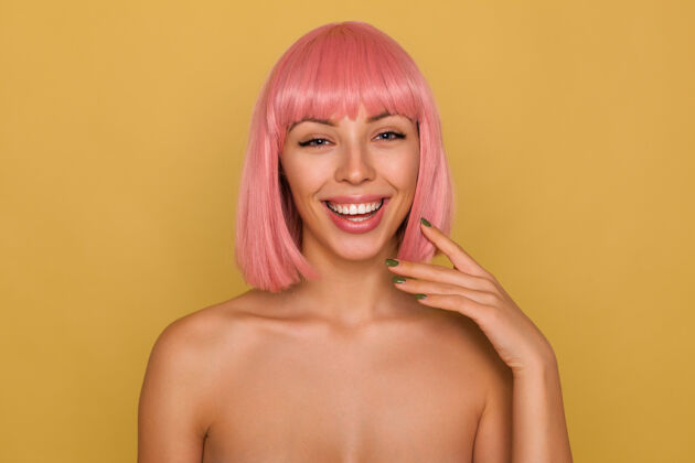 肖像快乐的年轻漂亮的粉红色头发的女人 留着鲍勃式的发型 笑容满面 手举着绿色的指甲贴在脸上 隔着芥末墙女人高兴姿势