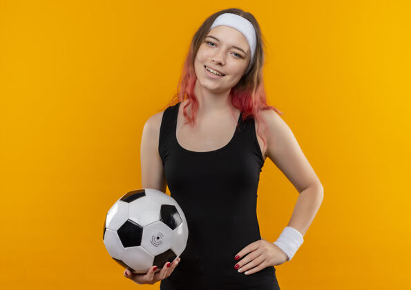 年轻身着运动服的年轻健身女士 手拿足球 脸上洋溢着幸福的笑容 站在橙色的墙上人看脸