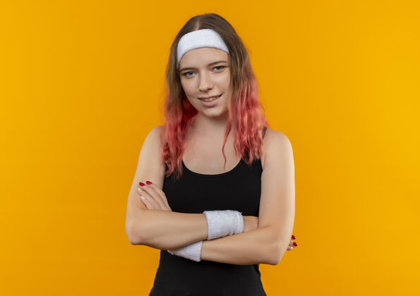 女人身穿运动服 双臂交叉放在胸前 脸上带着自信微笑的年轻健身女士站在橙色的墙上人市动
