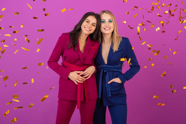 节日两位魅力四射的女士在紫罗兰色的墙上欢庆新年 穿着紫色和蓝色的时尚多彩的晚礼服 朋友们在一起玩得很开心 时尚潮流 金色的五彩纸屑派对气氛人人华丽