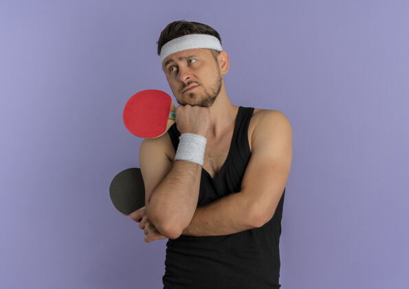 桌子一个戴着头巾的年轻人拿着两个乒乓球拍站在紫色的墙上 神情沉思地看着一边旁白男运动员