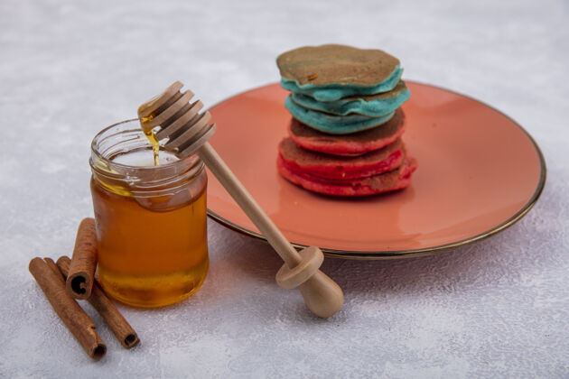 盘子侧视图蜂蜜与木勺肉桂和五颜六色的薄煎饼在一个橙色的盘子上白色的背景罐子肉桂甜味