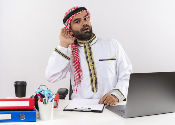 桌子身着传统服装的阿拉伯商人坐在桌子旁 手拿笔记本电脑 靠近耳朵 试图聆听办公室里的工作穿拿传统