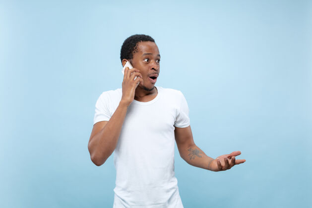 非洲身穿白衬衫 蓝色背景的非洲裔美国年轻人的半身特写肖像人类情感 面部表情 广告概念打电话 拿着智能手机手成人工作