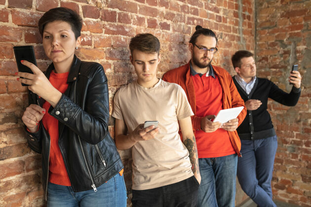 男孩一群快乐的白人年轻人站在砖墙后面分享来自智能手机或平板电脑的新闻 照片或视频 玩游戏 享受乐趣社交媒体 现代科技互联网观看连接