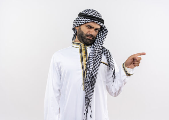 穿一个穿着传统服装的阿拉伯男人 皱着眉头 手指着站在白墙上的一边看传统指