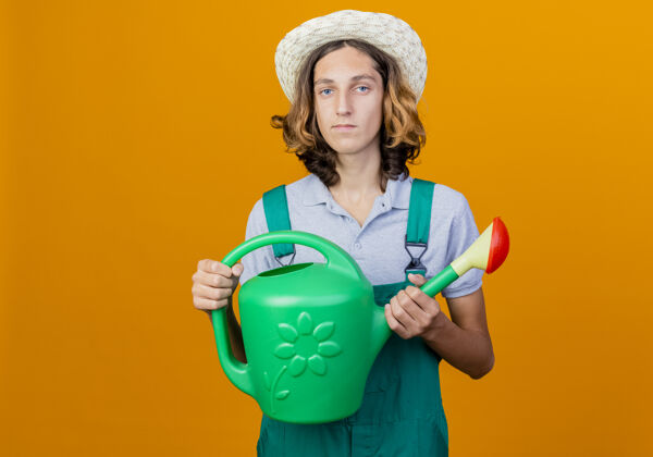 脸年轻的园丁穿着连体衣戴着帽子拿着浇水罐严肃年轻男人