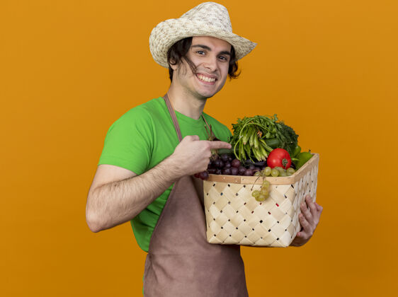 帽子年轻的园丁 围着围裙 戴着帽子 手里拿着装满蔬菜的箱子 看着摄像机 兴高采烈地笑着 用食指指着站在橙色背景上的箱子年轻指向欢呼