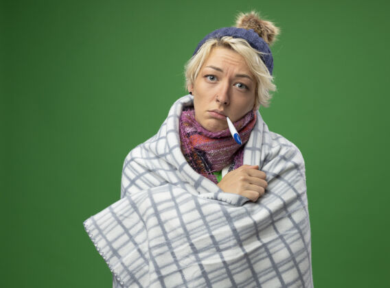 头发有病的不健康的女人 短头发 戴着暖和的围巾 帽子 裹着毯子 嘴里叼着温度计 温度在绿色背景上帽子围巾体温计