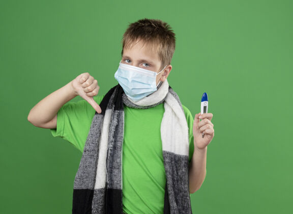 周围生病的小男孩穿着绿色t恤 脖子上围着暖和的围巾 戴着护面面具 手拿着温度计 站在绿色背景上竖起大拇指温暖向下疾病