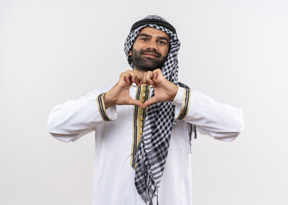 阿拉伯语身着传统服装的阿拉伯男子站在白墙上做着心形手势 手指微笑着 快乐而积极男人积极微笑