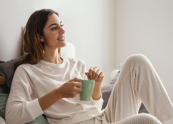休闲笑脸女人在床上喝咖啡的侧视图休假室内女人
