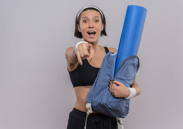 人穿着运动服的年轻健身女士拿着背着瑜伽垫的背包 兴高采烈地指着白墙上的摄像机目录姿势运动装