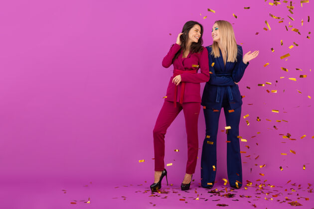 乐趣两位魅力四射的女士在紫罗兰色的墙上欢庆新年 穿着紫色和蓝色的时尚多彩的晚礼服 朋友们在一起玩得很开心 时尚潮流 金色的五彩纸屑派对气氛五彩纸屑美丽女人