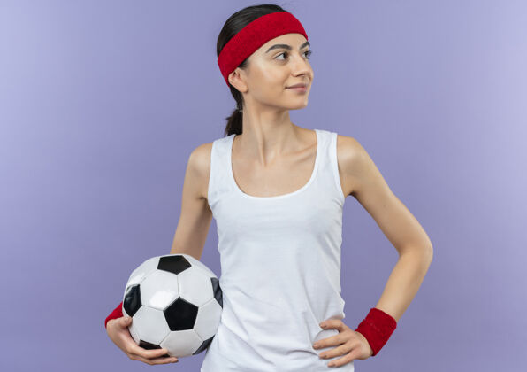 姿势身穿运动服 头箍 手持足球 脸上带着自信微笑的年轻健身女士站在紫色的墙上健康抱足球