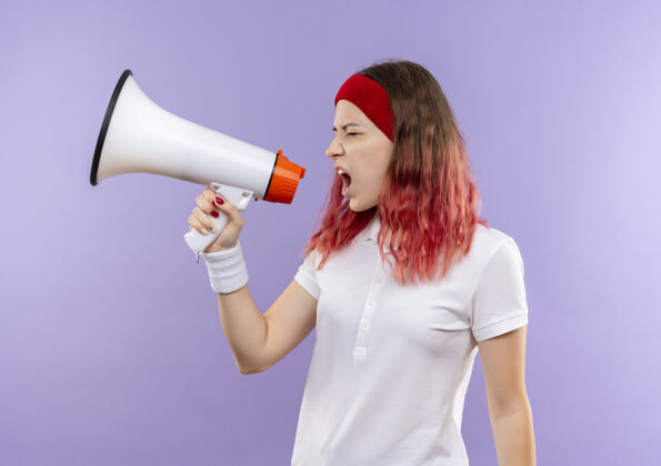 人年轻的运动型女人站在紫色的墙上对着扩音器大喊大叫 表情咄咄逼人侵略性喊叫立场