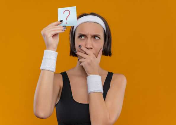 沉思戴着头带的运动装的年轻健身女士拿着带问号的提醒纸 带着沉思的表情看着它 想着站在橙色的墙上问题纸人