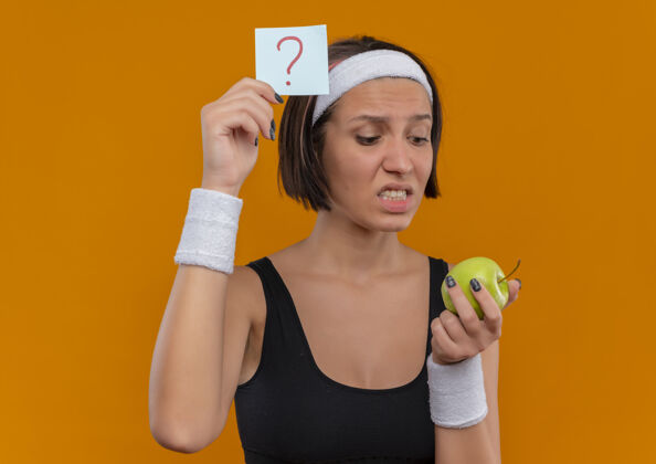 困惑身穿运动服的年轻健身女士 头上戴着带问号的提醒纸 手里拿着绿色的苹果 站在橙色的墙上困惑而焦急地看着它健身运动员运动