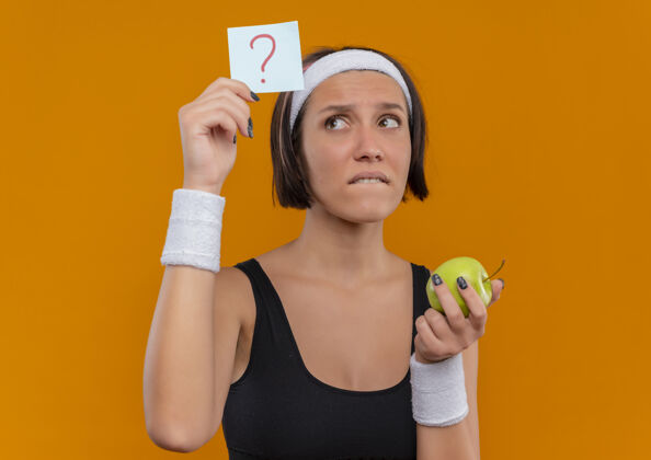 姿势穿着运动服的年轻健身女士 头上戴着带问号的提醒纸 手里拿着绿色的苹果 站在橙色的墙上困惑地看着纸困惑女人纸