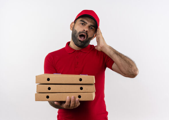 男人留着胡须的送货员穿着红色制服 戴着帽子 手里拿着一叠披萨盒 站在白墙上张大嘴巴 既震惊又困惑胡子送货拿着