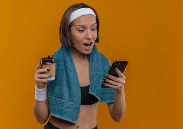 运动一位身穿运动服 脖子上套着毛巾 手里拿着咖啡杯 看着智能手机屏幕的年轻健身女士站在橙色的墙上 既惊讶又高兴健康看站