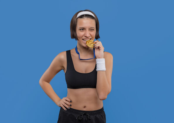 健身身穿运动服 脖子上挂着金牌的年轻健身女士站在蓝色的墙上 咬牙切齿地微笑着女性周围运动员