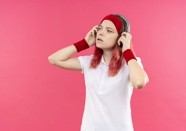 人物戴着头戴式耳机的年轻运动女性站在粉色的墙上一边听音乐一边看运动女性站立