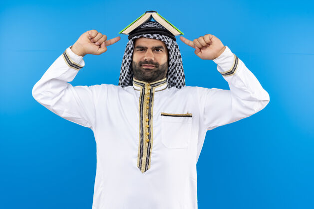 头身着传统服装的阿拉伯商人 头上戴着一本书 手指指着书 微笑着站在蓝色的墙上传统微笑站
