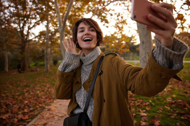 白天快乐的年轻迷人的棕色头发的女性穿着温暖舒适的衣服 举起她的手 同时自拍她的smarthope 站在模糊的公园黑发化妆品理发