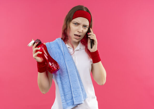 运动戴着头巾 肩上扛着毛巾 手里拿着一瓶水的年轻运动女郎站在粉红色的墙上 一边讲电话 一边显得困惑和焦虑水运动员瓶子