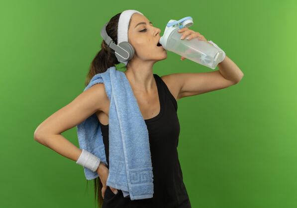 姿势穿着运动服的年轻健身女士 头上戴着头巾 肩上扛着毛巾 锻炼后站在绿墙边喝水饮水人运动
