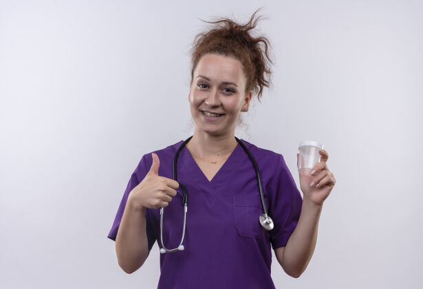 女人年轻的女医生穿着医疗制服 手持听诊器 拿着测试罐 面带微笑 竖起大拇指站在白色的墙上年轻人测试抱着