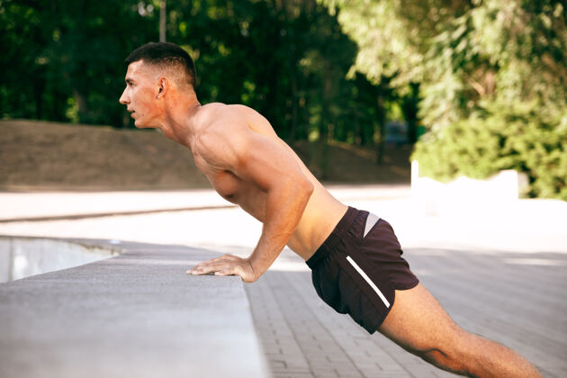 运动员一个肌肉发达的男运动员在公园里锻炼体操 训练 健身锻炼 柔韧性夏日城市在阳光明媚的日子背景场上积极健康的生活方式 青春 健美伸展运动健美