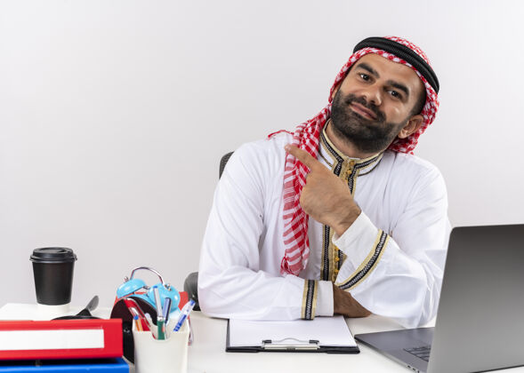 手指身着传统服装的阿拉伯商人坐在桌旁 用食指指着左边的笔记本电脑 看上去自信地在办公室工作电脑左笔记本电脑
