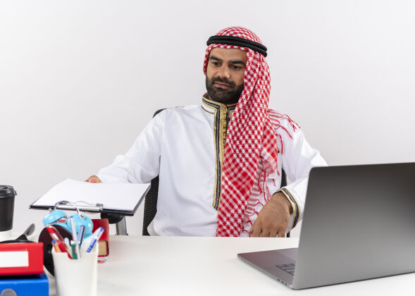 笔记本电脑身着传统服装的阿拉伯商人坐在桌旁 手提电脑拿着剪贴板 空白页在办公室工作坐着持有阿拉伯语