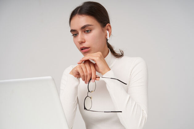 女士一个年轻的蓝眼睛的黑发女人的肖像 扎着马尾辫 困惑地看着笔记本电脑的屏幕 一边戴着眼镜 一边在白墙上摆姿势欧洲困惑笔记本电脑
