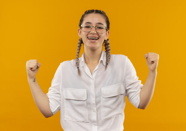 快乐戴着眼镜 梳着辫子 穿着白衬衫的年轻女学生握紧拳头 高兴而兴奋地站在橙色的墙上为自己的成功而欢呼姿势学生衬衫