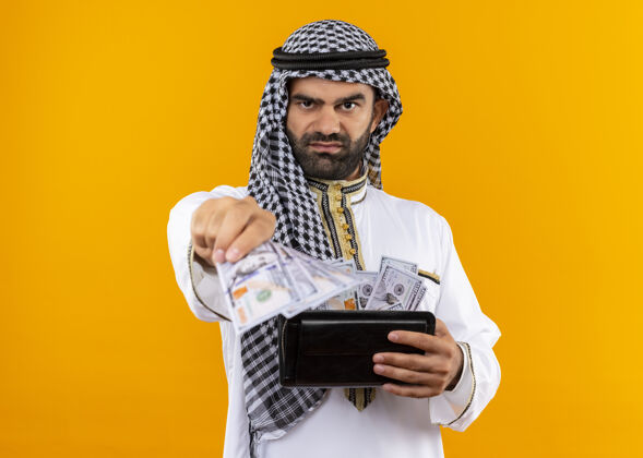 传统身着传统服装的阿拉伯商人手持钱包 面带严肃表情 站在橙色的墙上站阿拉伯语商务人士