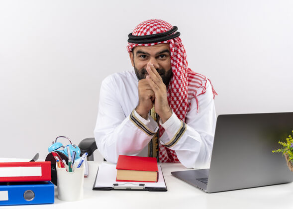 微笑身着传统服装的阿拉伯商人坐在桌旁 带着笔记本电脑 狡黠地笑着 在办公室里工作愉快穿着传统电脑