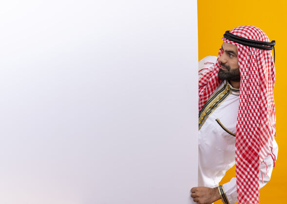 严重身着传统服装的阿拉伯商人从空白的广告牌中探出眼睛 严肃地看着橙色墙壁上的广告牌阿拉伯语空白脸