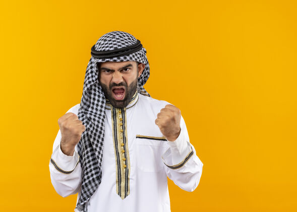 穿身着传统服装的阿拉伯商人站在橙色的墙上怒气冲冲地握紧拳头商人脸握紧