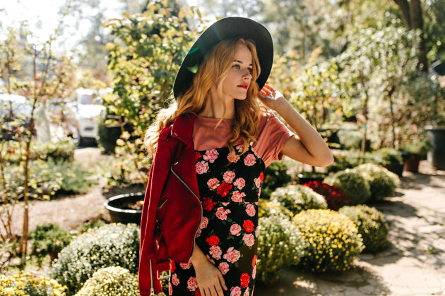 开花迷人的卷发女子 头戴宽边帽 身穿红色皮夹克 黑色印花连衣裙 若有所思地遥望远方 在花园里享受春日金发肖像花店