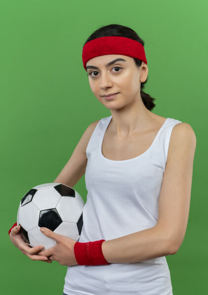 表情身穿运动服 头箍 手持足球 表情自信的年轻健身女孩站在绿色的墙上女孩健身运动装